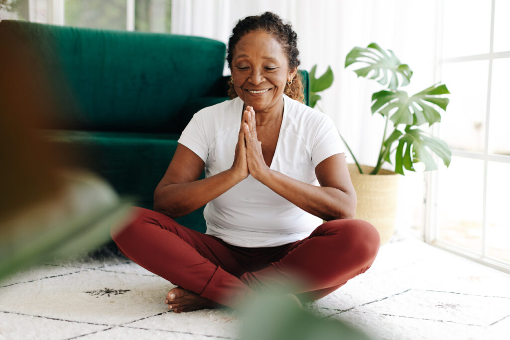 Senior woman improving mental wellbeing through yoga, meditating at home with a serene smile. Elderly woman doing a regular yoga practice to support her health, reduce stress and promote inner peace.