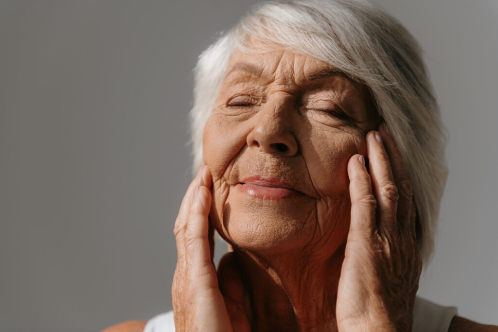 Portrait of beautiful senior woman touching face and keeping eyes closed against grey background, defining the beauty of aging